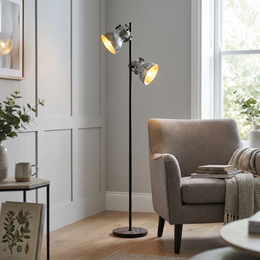 Industrial Eglo Barnstaple floor lamp with two zinc shades and a wooden base standing beside a grey armchair.