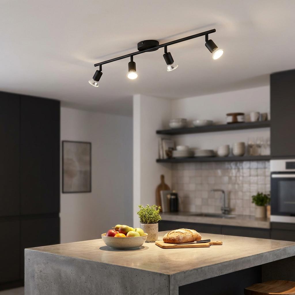 A black four-light track spotlight mounted on a white kitchen ceiling, illuminating a modern concrete island.