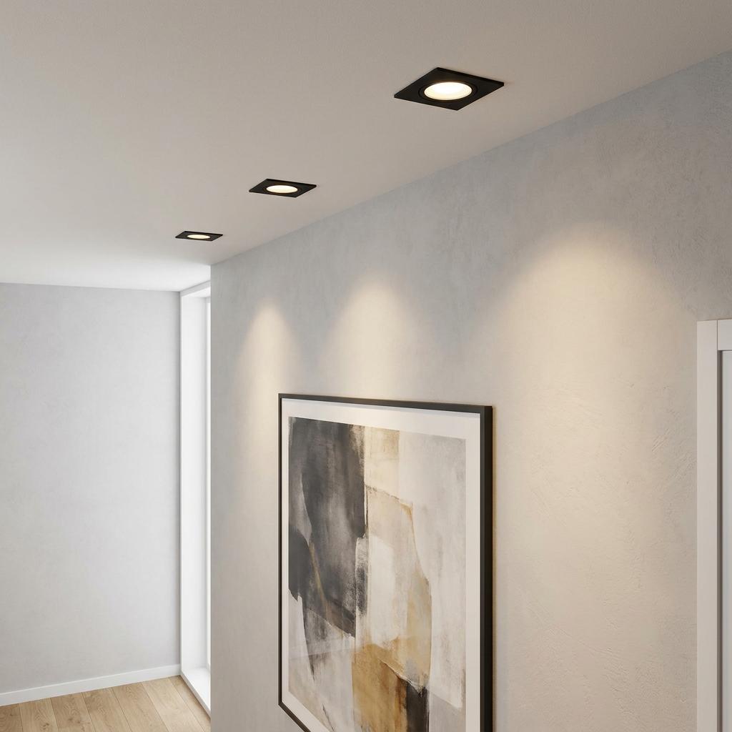Three black square recessed spotlights on a ceiling illuminating an abstract painting on a light wall.