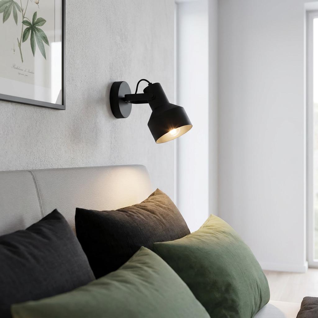 Black industrial wall spotlight illuminated above a bed with grey and green pillows.
