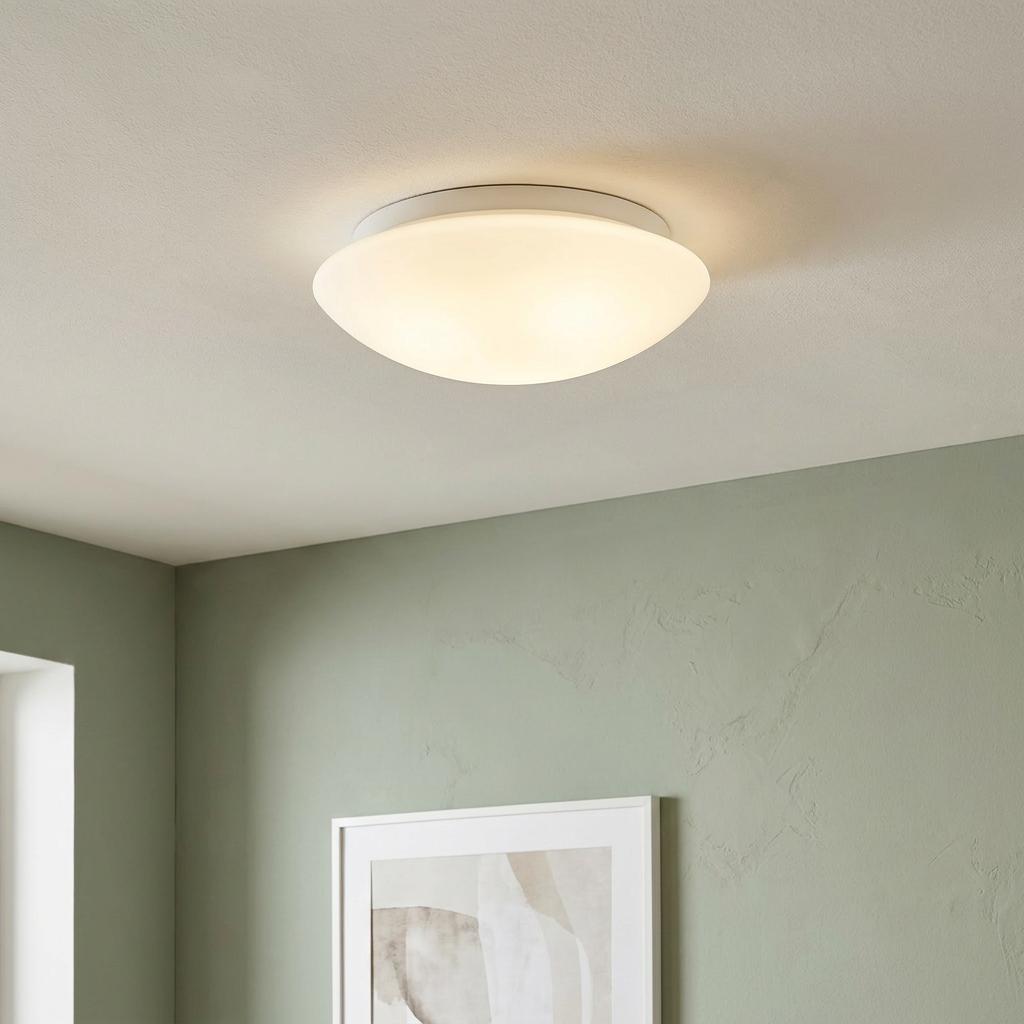 White opal glass flush ceiling light illuminated on a white ceiling above a sage green wall.