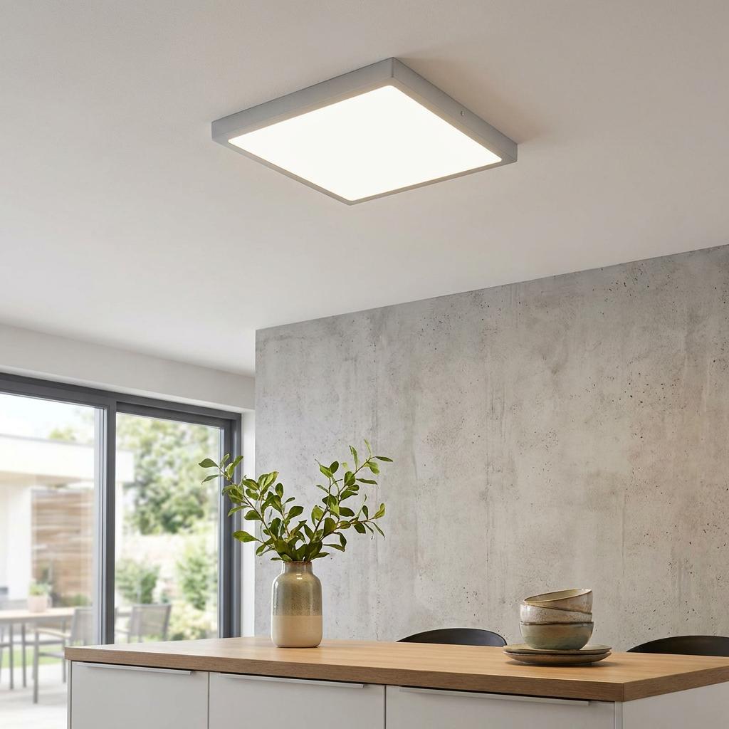 Square silver flush LED ceiling light illuminating a modern room with a concrete wall and wooden table.
