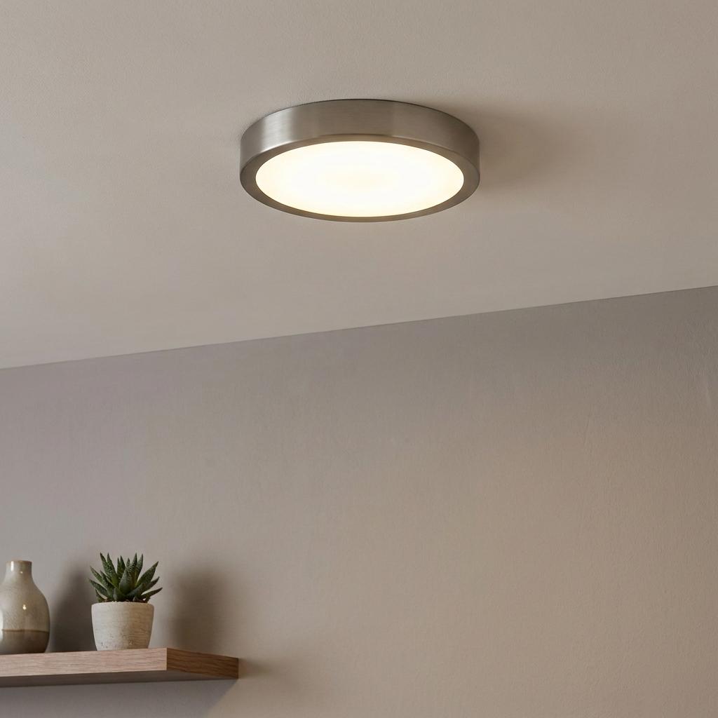Round nickel flush mount LED ceiling light illuminating a neutral-toned room above a wooden shelf.