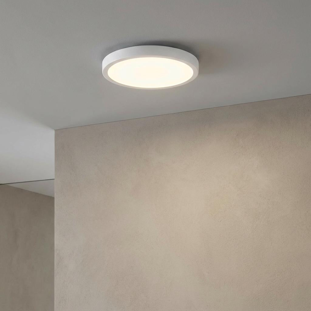 Round white LED flush ceiling light mounted on a white ceiling next to a neutral textured wall.