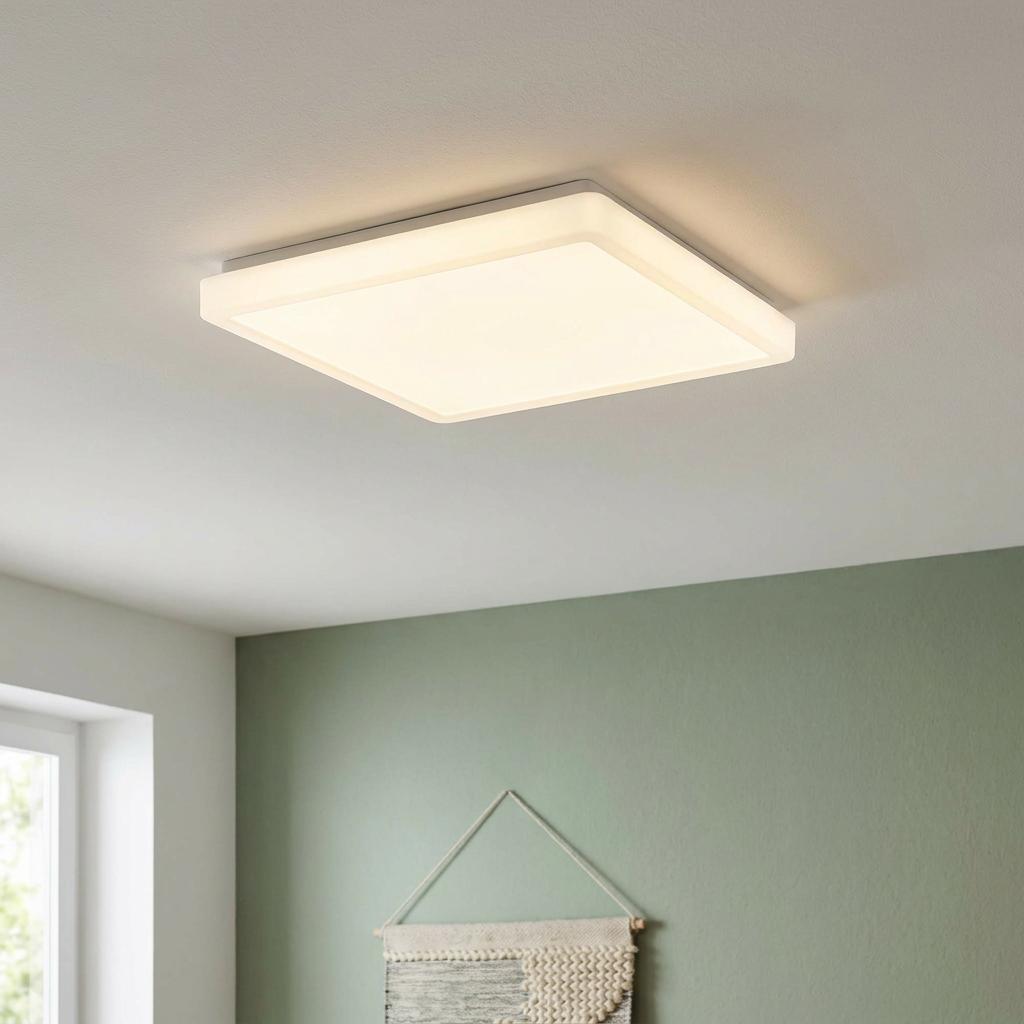 Square white LED flush ceiling light glowing with warm light on a white ceiling above a green wall.