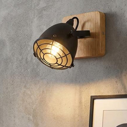 Industrial black spotlight with a wire cage and wooden base, mounted on a textured grey wall in a living room.