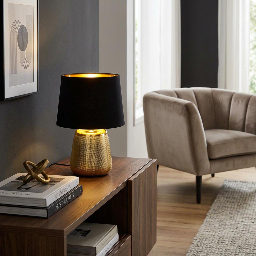 Eglo Manalba 1 gold ceramic table lamp with a black fabric shade on a wooden cabinet in a contemporary living room.