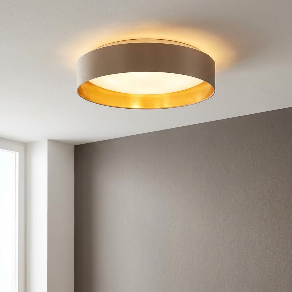Round taupe fabric flush ceiling light with a gold interior glowing on a white ceiling above a brown wall.