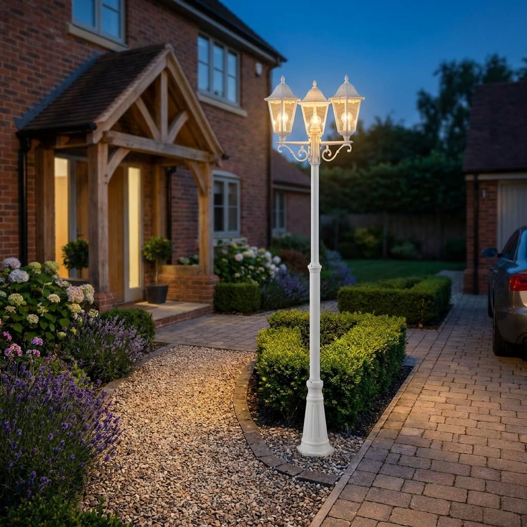 White three-head lantern lamp post illuminating a garden pathway in front of a brick house at dusk.