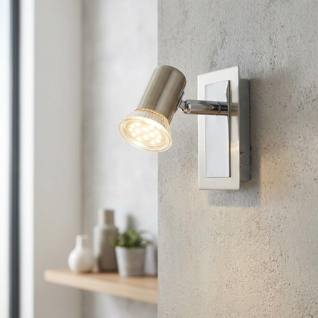 Satin nickel wall spotlight with an illuminated bulb mounted on a textured grey wall.