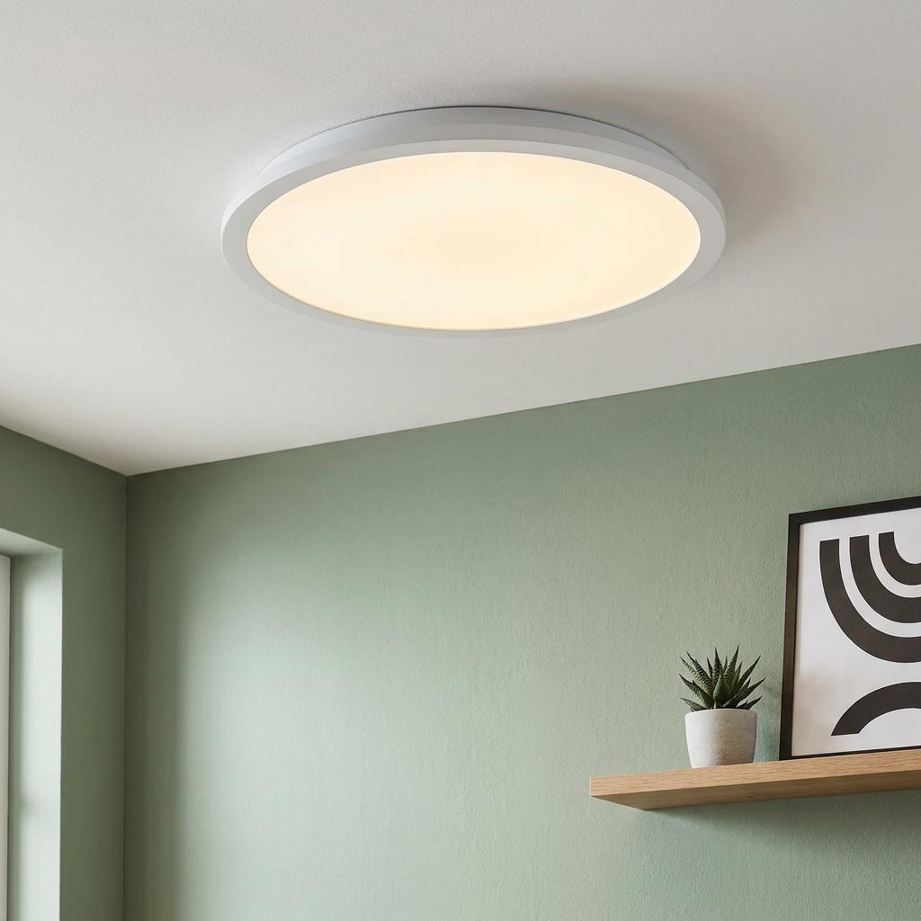 Round white LED flush ceiling light illuminated in a room with a sage green wall and wooden shelf.