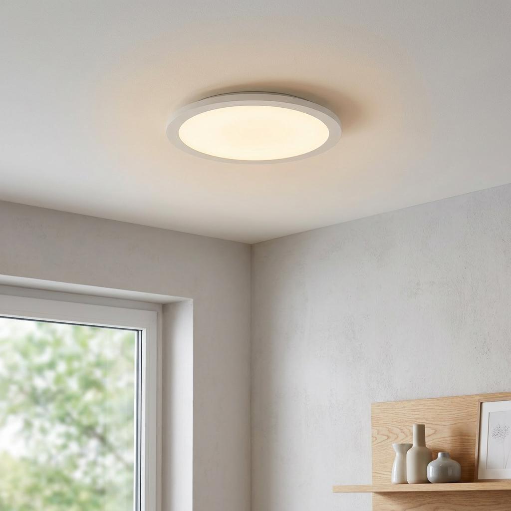 Round white LED ceiling flush light illuminated in a neutral room with a window and decorative shelf.