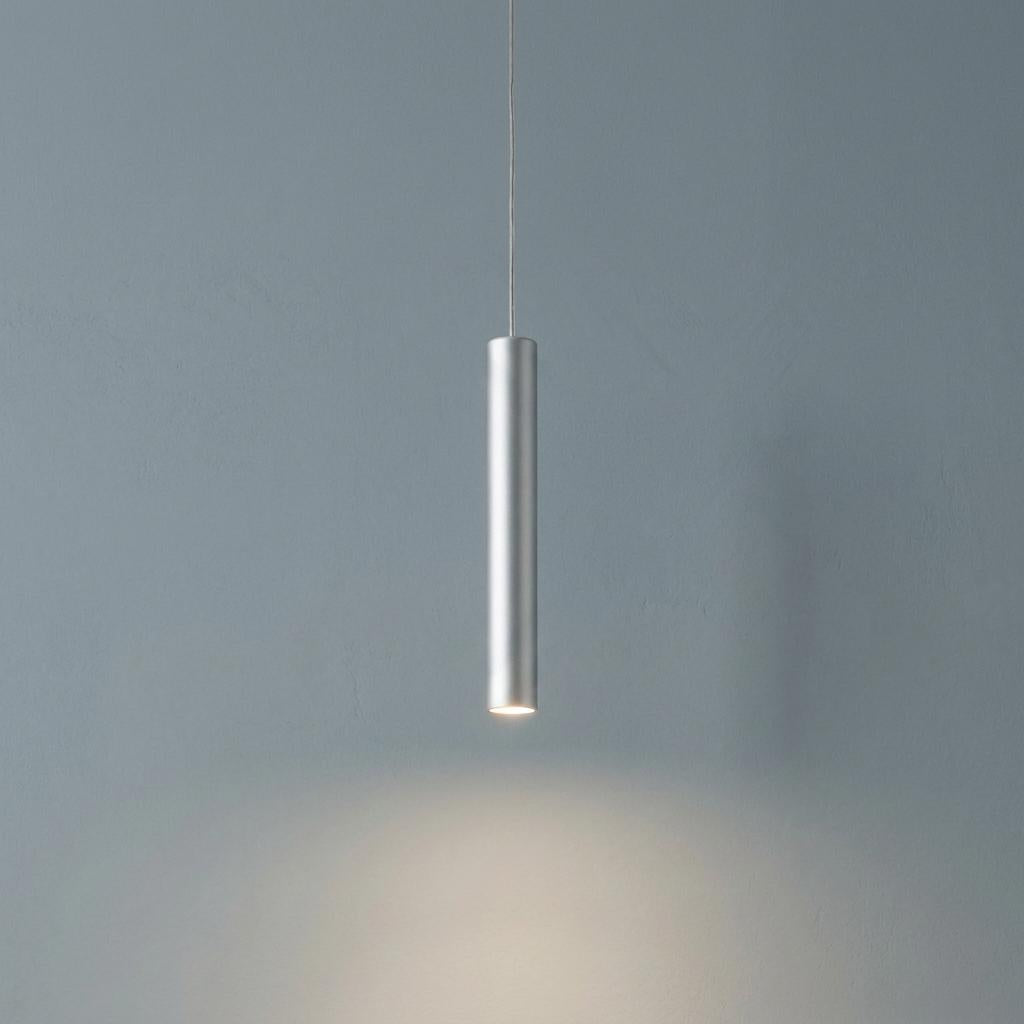 Sleek silver cylindrical LED pendant light hanging against a grey textured wall with a downward glow.