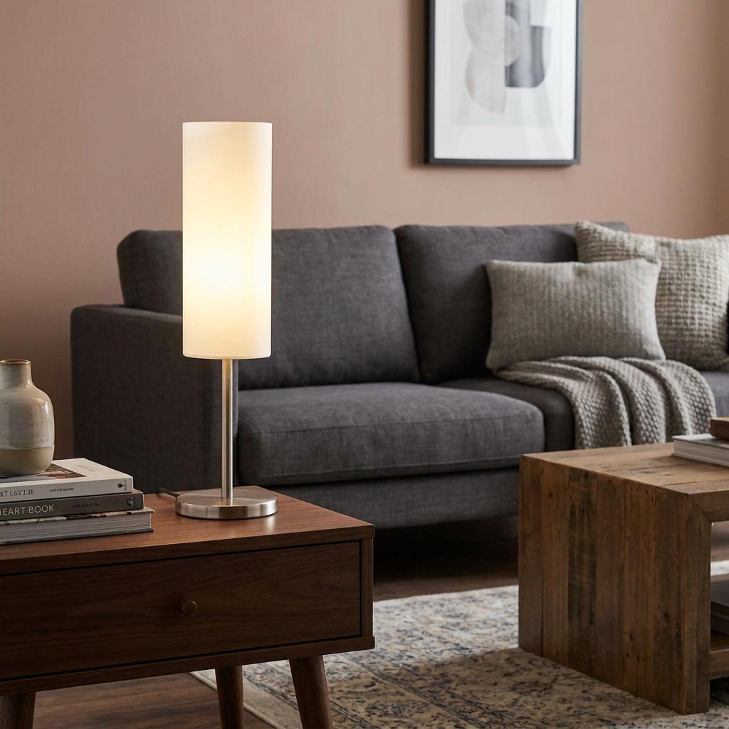 Illuminated cylindrical white glass table lamp with a brushed nickel base on a wooden side table in a living room.