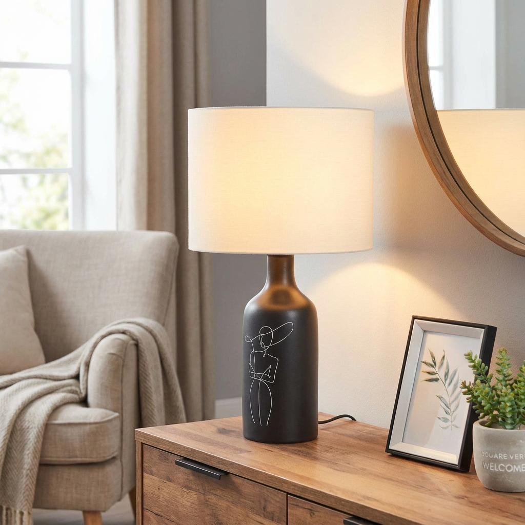 Lit black ceramic table lamp with a line-art woman illustration and white drum shade on a wooden sideboard.
