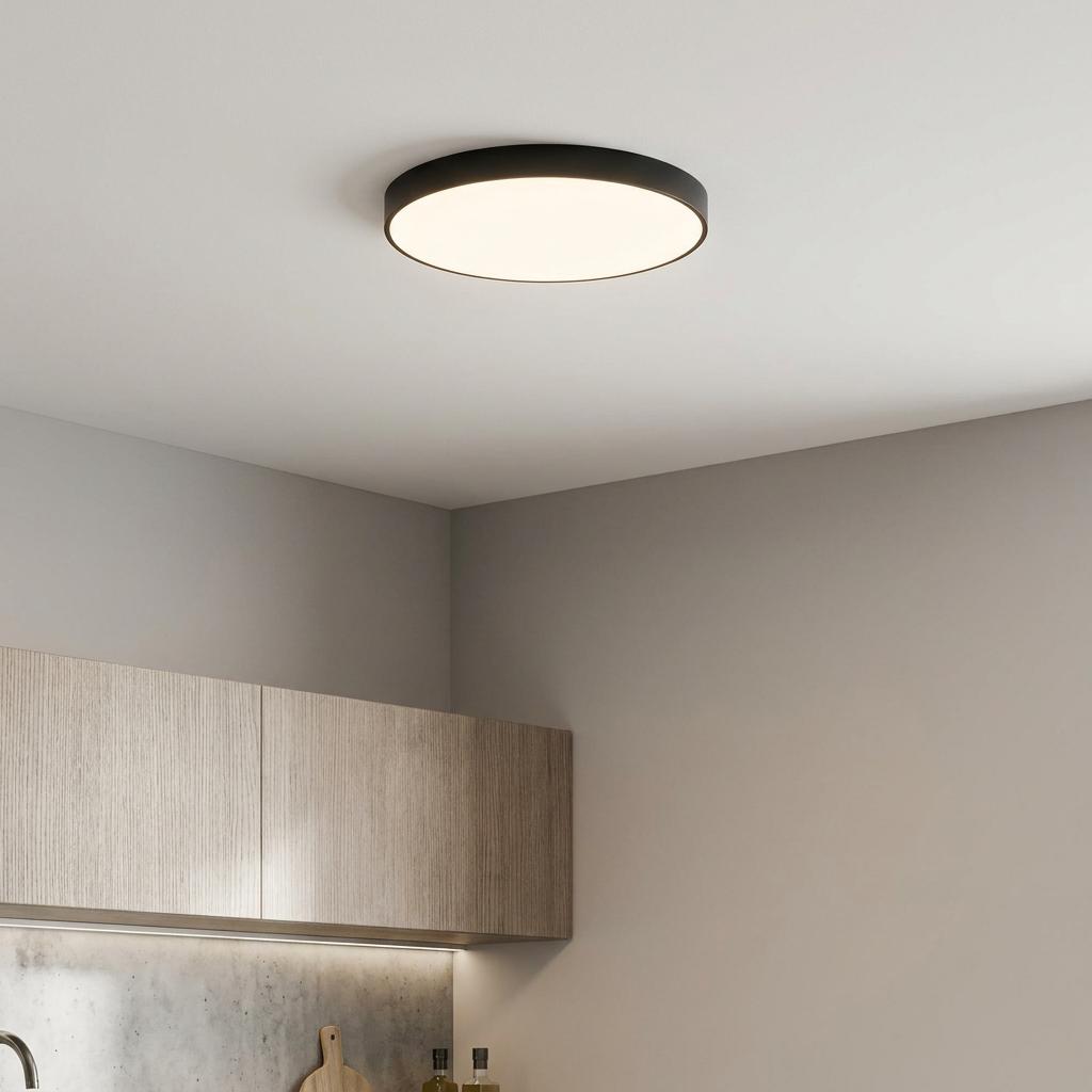 A modern black round flush ceiling light illuminating a minimalist kitchen with wooden cabinets.