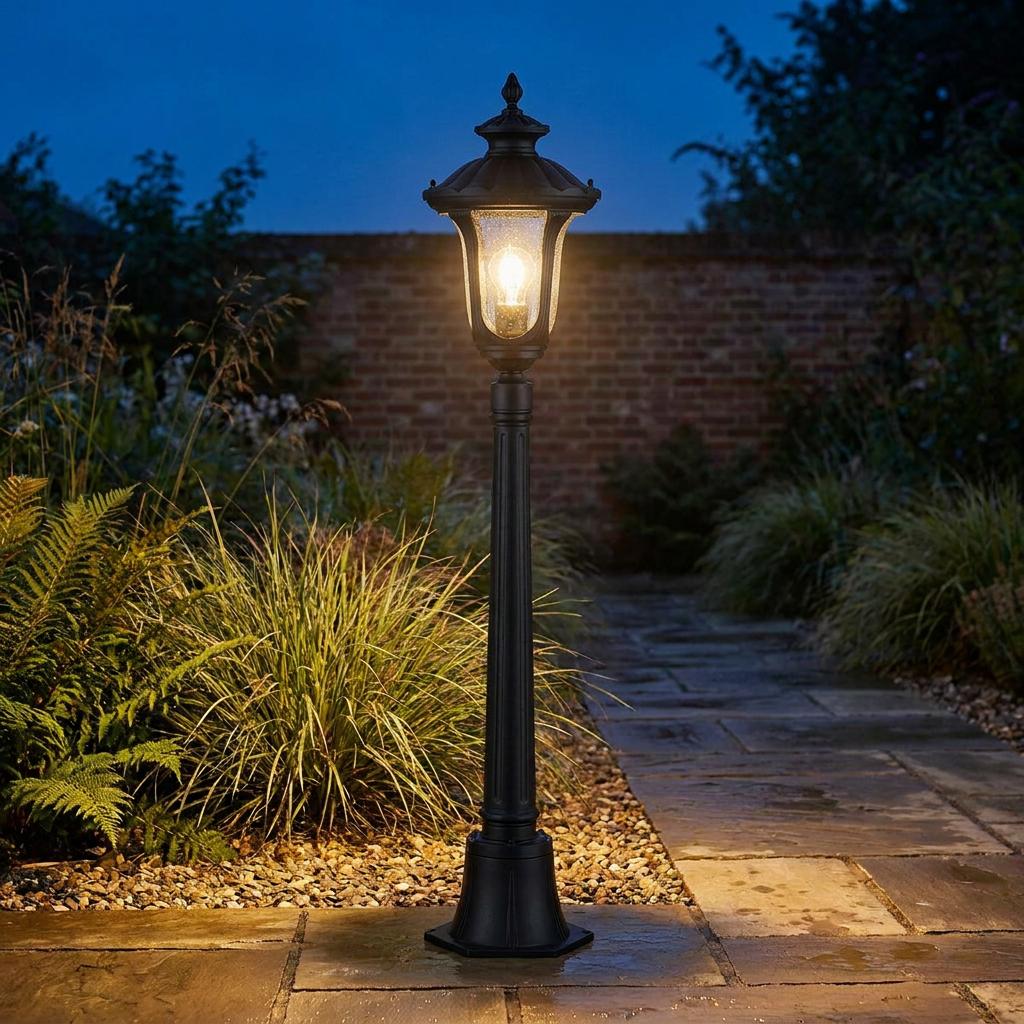 A black Elstead Chicago outdoor bollard light with a warm glow illuminates a paved garden path at dusk.