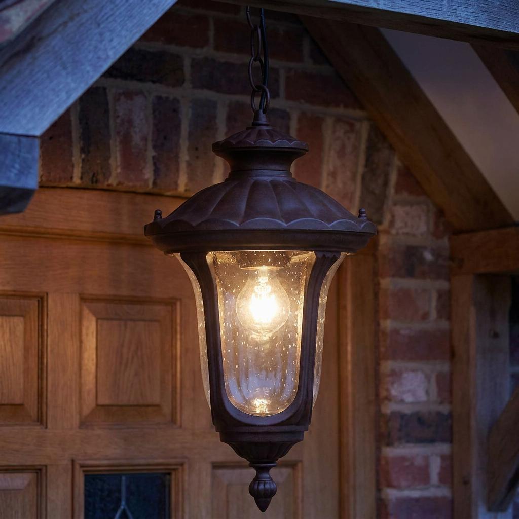 A rusty bronze patina outdoor lantern with seeded glass hangs illuminated against a wooden door and brick wall.
