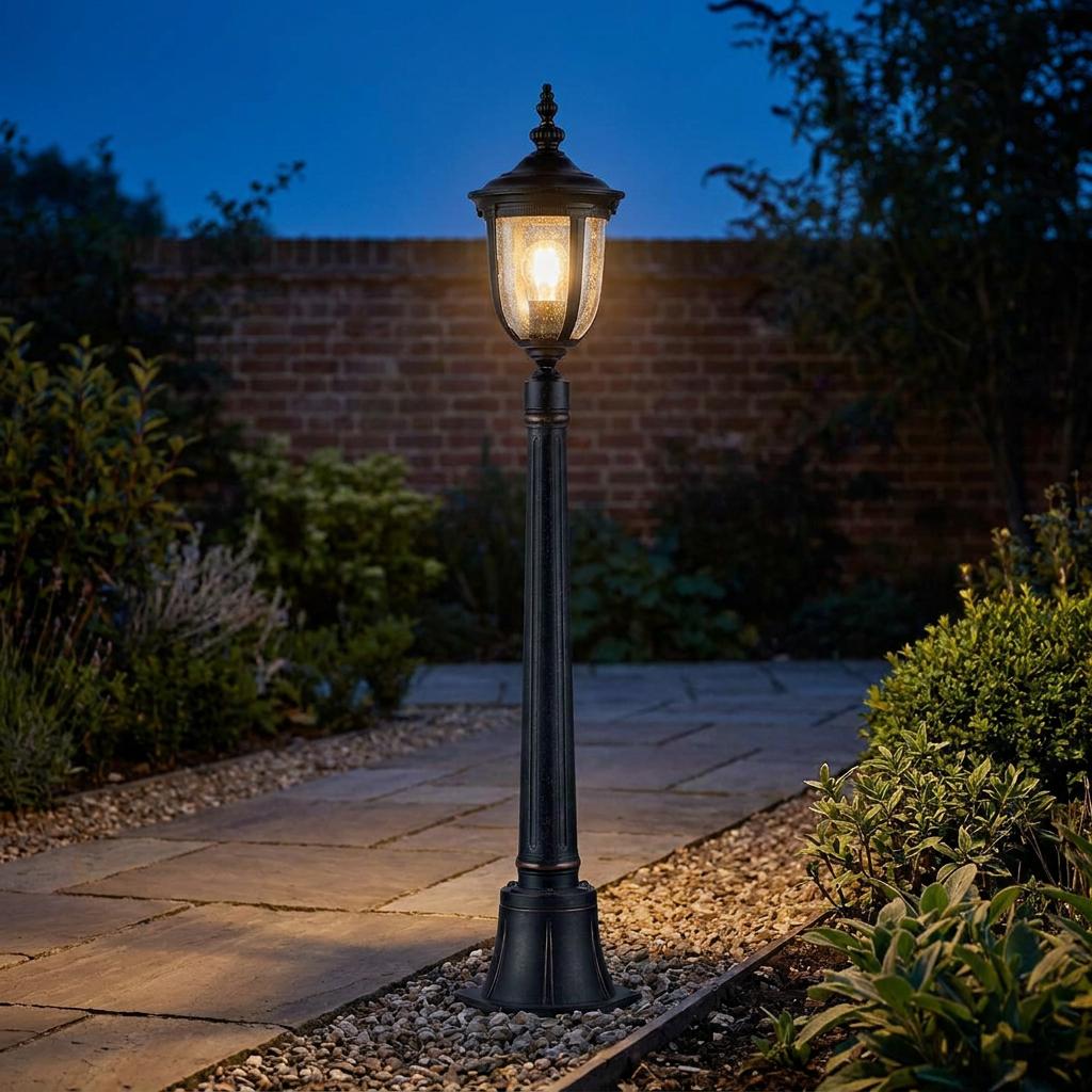 Traditional black outdoor bollard light with seeded glass lantern glowing on a stone garden path at twilight.