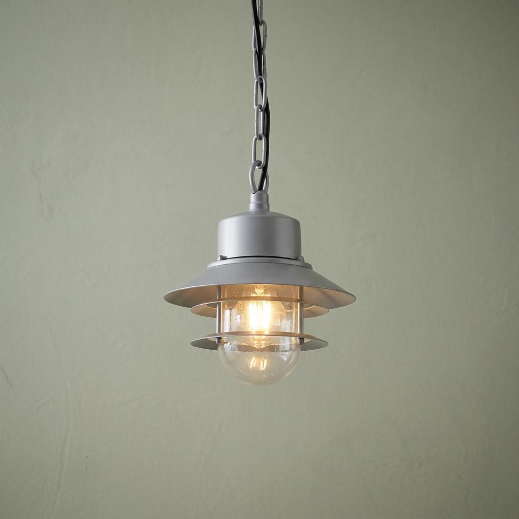 Silver outdoor chain lantern with a lit filament bulb hanging against a textured sage green wall.