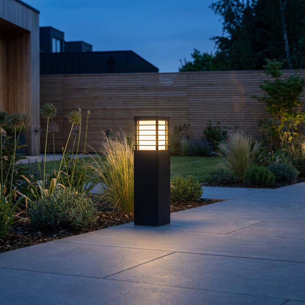 Modern black bollard light with a warm glow illuminating a garden path and flower beds at twilight.