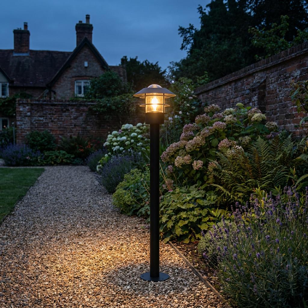 A black outdoor bollard light illuminates a gravel garden path at twilight, surrounded by flowers and greenery.