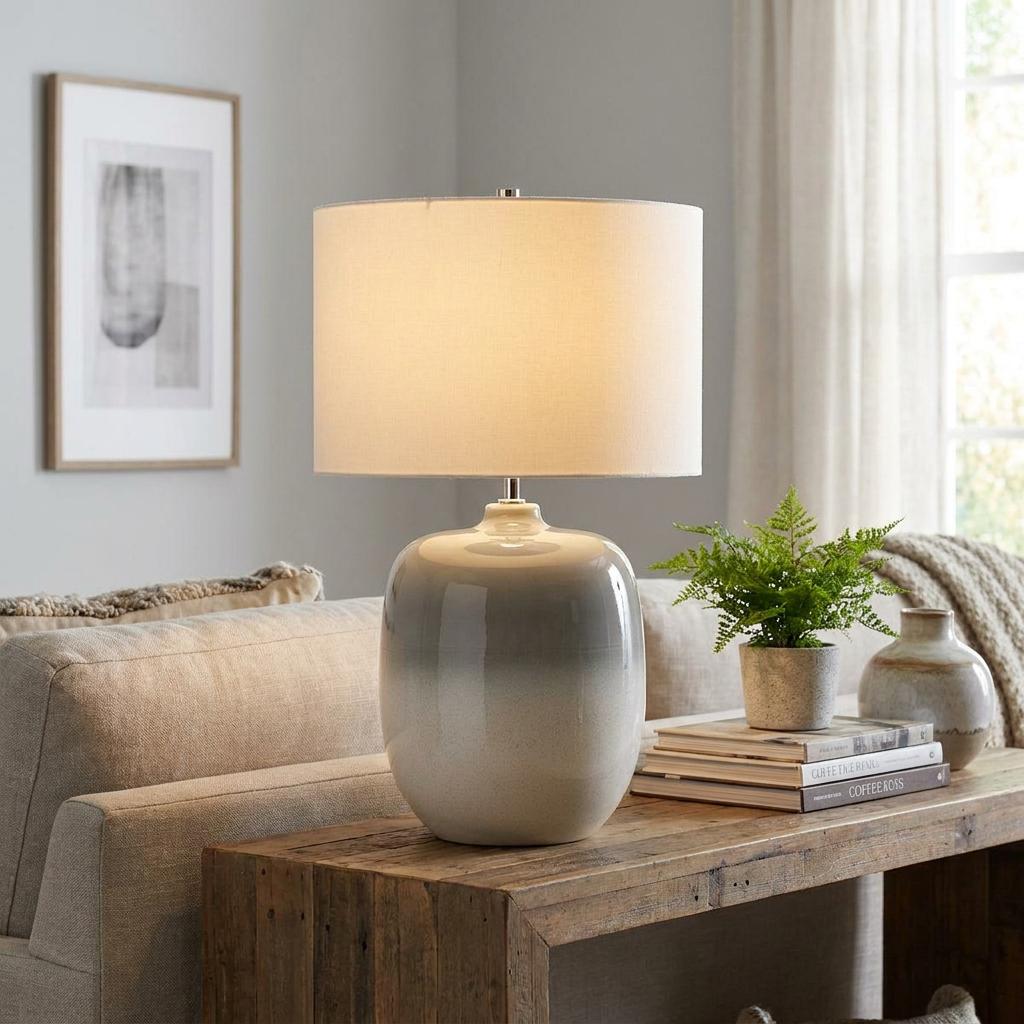 Lit ceramic table lamp with a grey and white ombre base and cream drum shade on a rustic wooden console table.