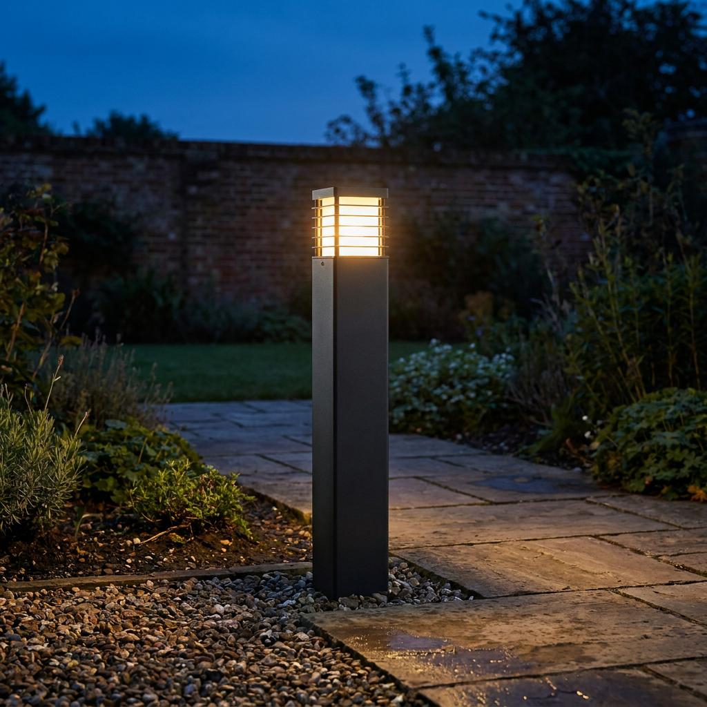 Elstead Lighting Halmstad black bollard light glowing on a stone path in a garden at dusk.