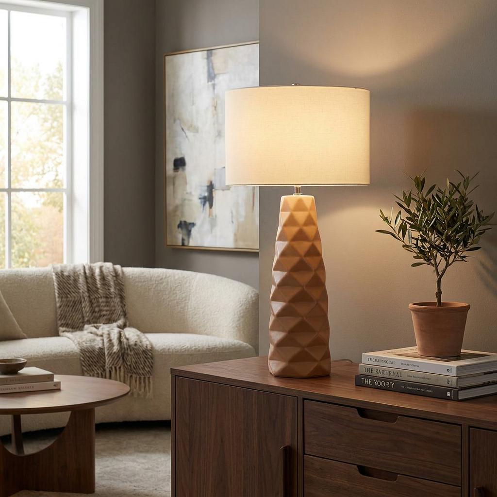 Caramel ceramic table lamp with a geometric base and drum shade on a wooden sideboard in a cozy living room.