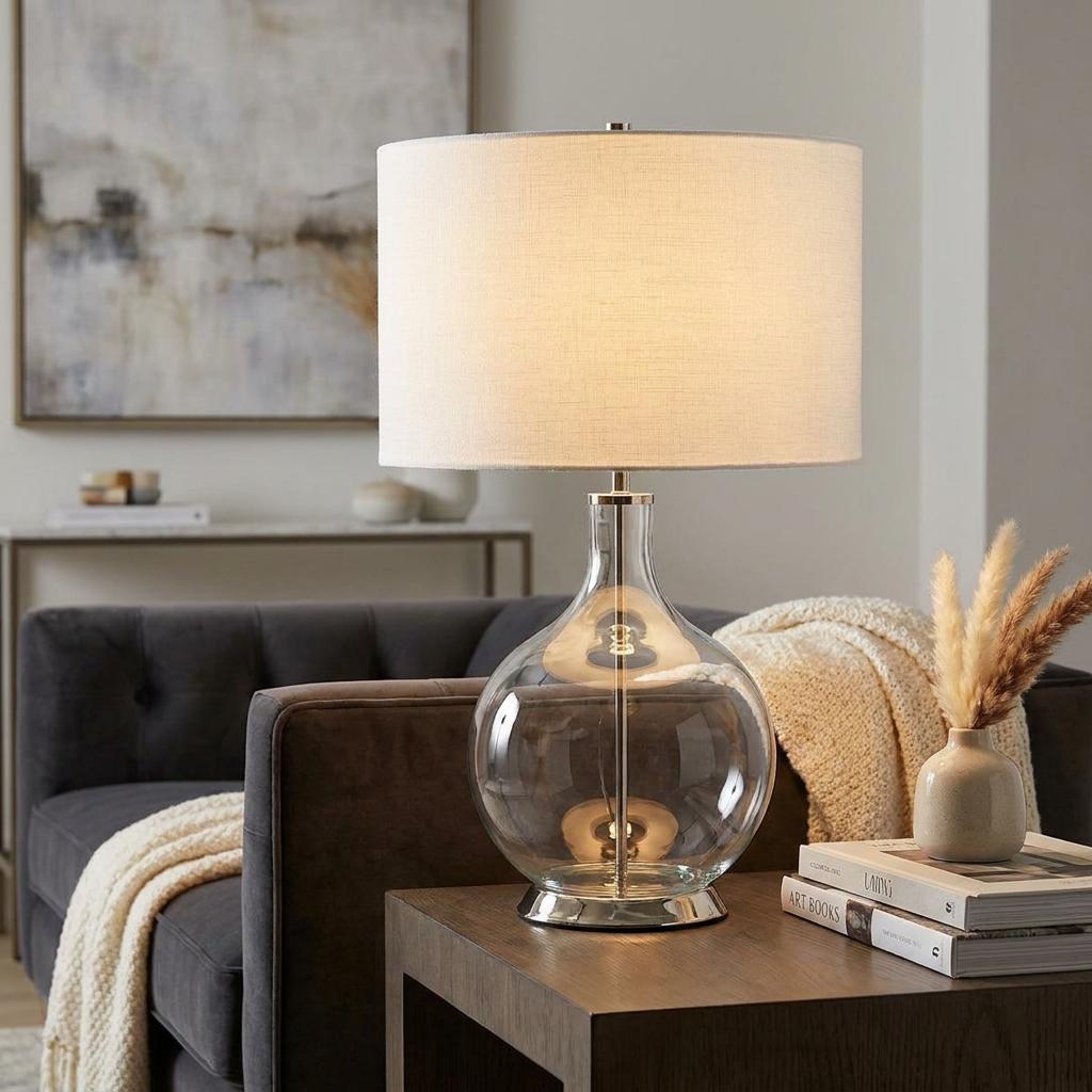 Elstead Orb table lamp with a clear glass base and white drum shade, lit on a side table in a modern living room.