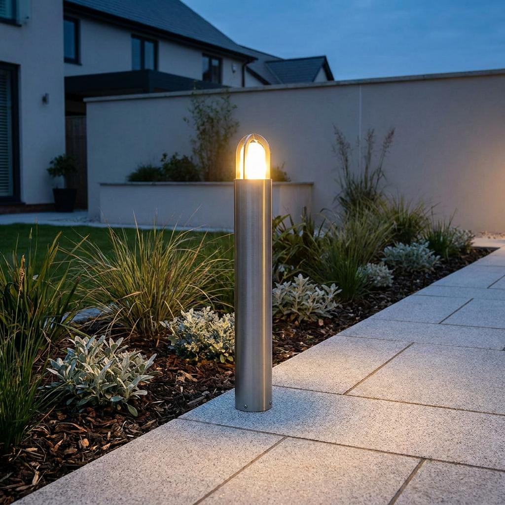 A brushed silver bollard light glowing warmly beside a paved garden walkway at dusk.