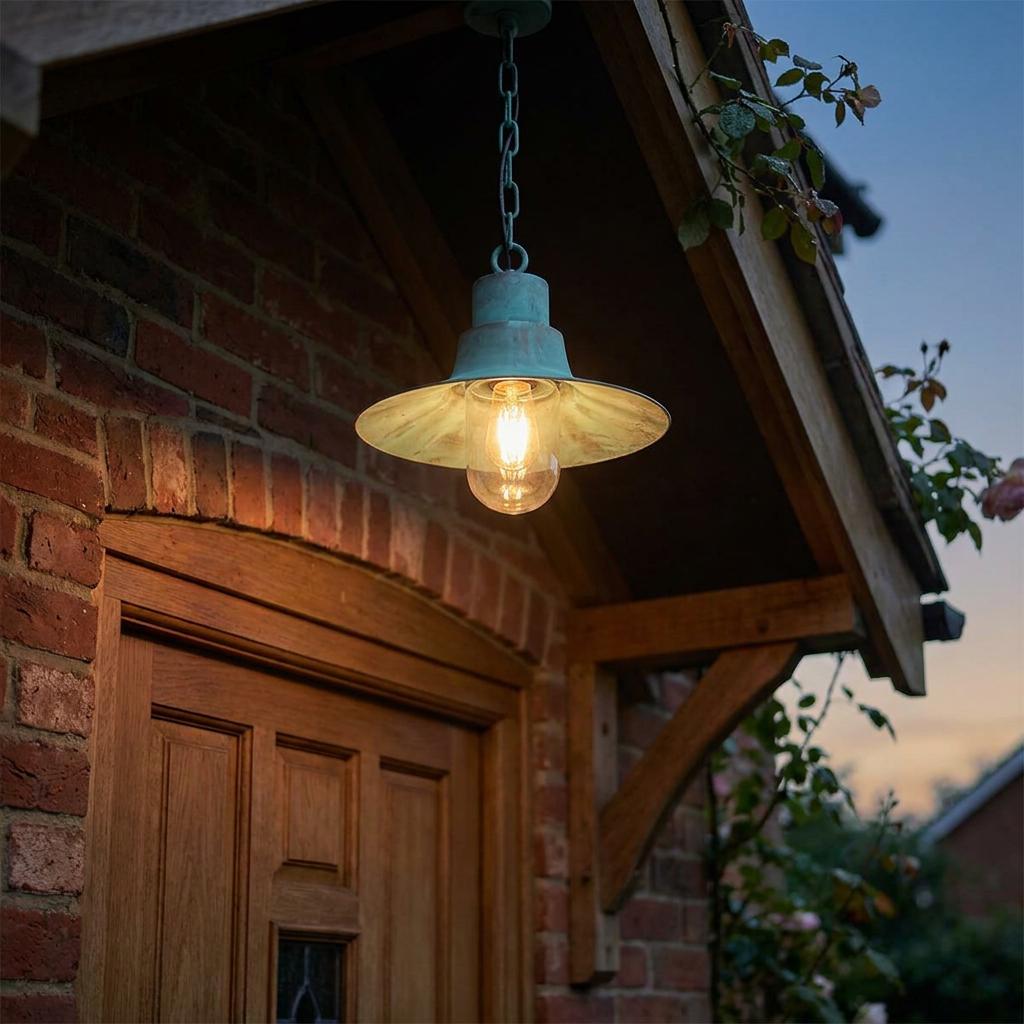 Verdigris outdoor pendant light with a glowing bulb hanging under a wooden porch outside a brick house.