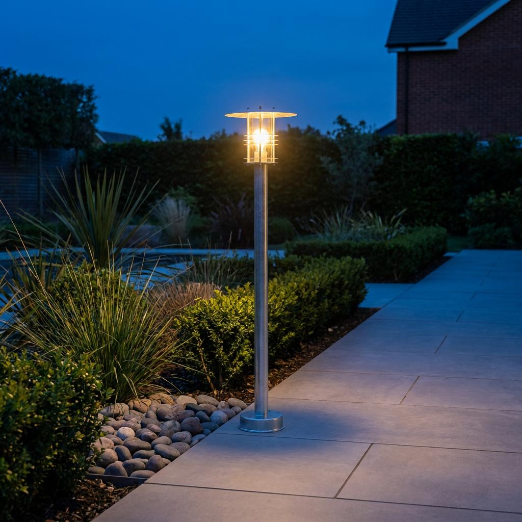 Warmly lit galvanised steel outdoor bollard light standing beside a paved garden path at dusk.