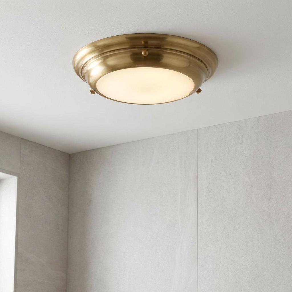 Aged brass flush mount LED ceiling light with a frosted glass diffuser on a white ceiling in a grey tiled bathroom.