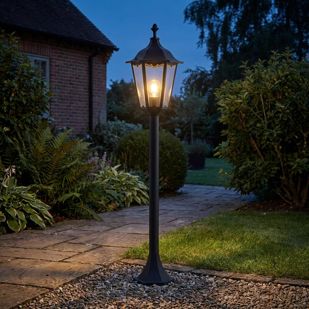 Lit Endon Burford black lantern bollard on a stone garden path at twilight with lush green foliage behind.