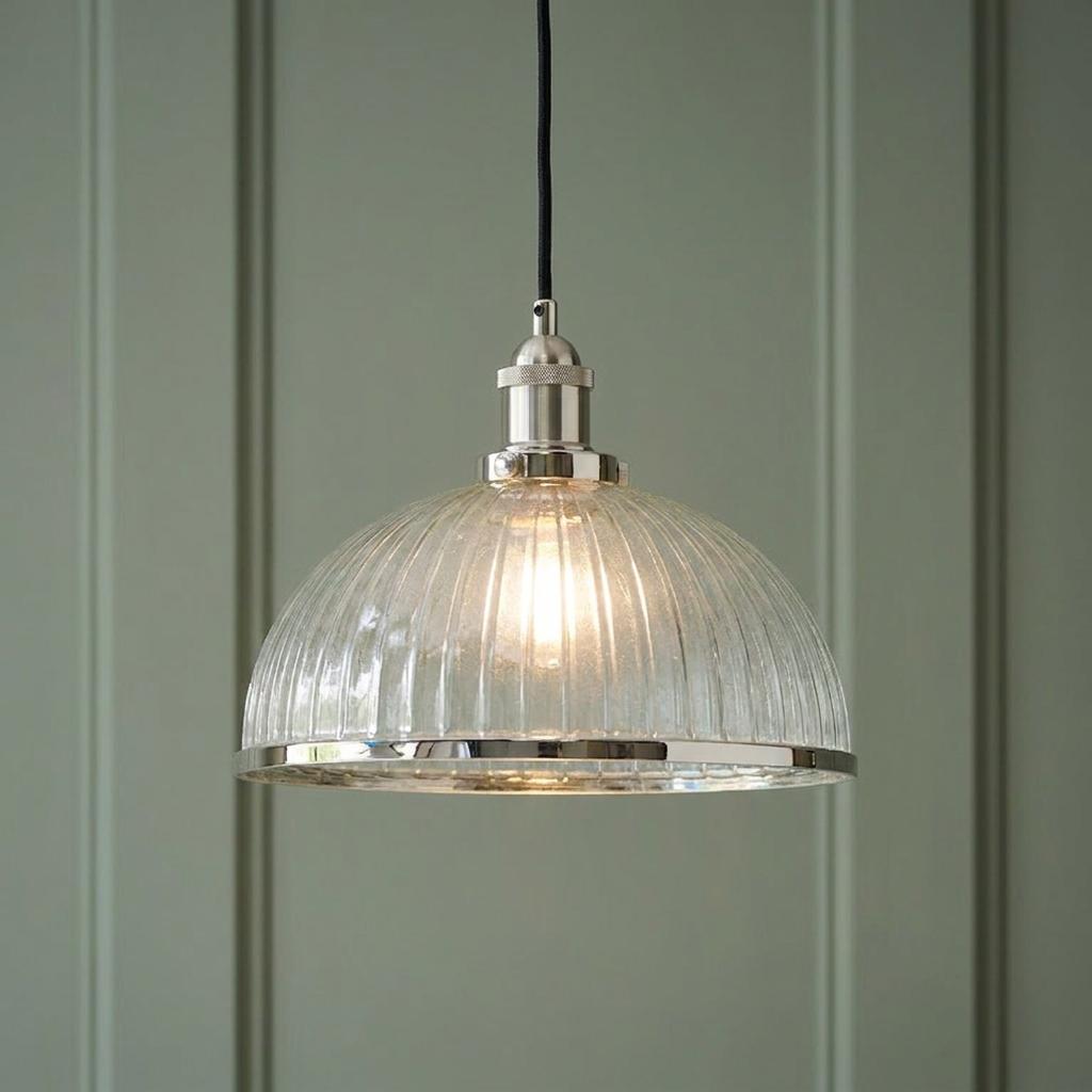 Lit industrial pendant light with a ribbed glass shade and polished nickel trim against a sage green paneled wall.