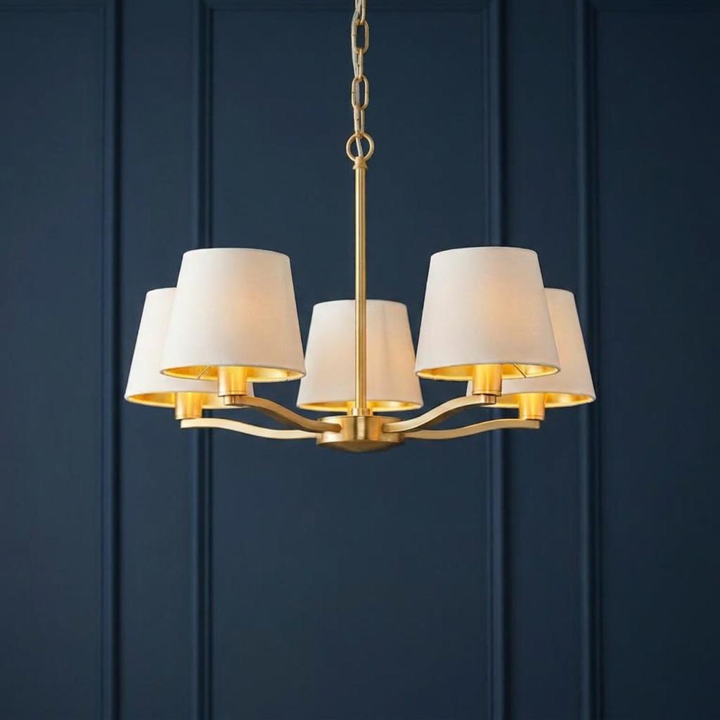 Five-light gold pendant chandelier with white fabric shades against a dark blue paneled wall.