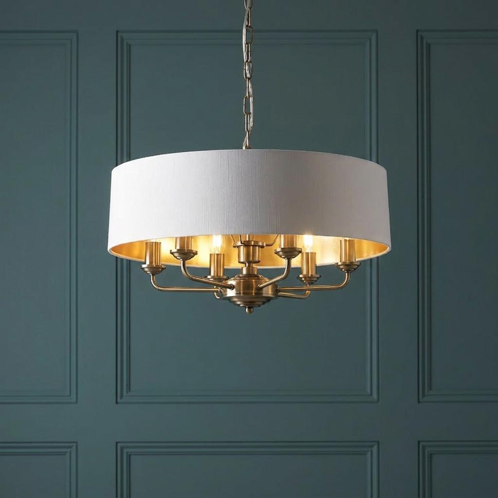 Antique brass 6-light pendant with a white drum shade and gold interior against a teal paneled wall.