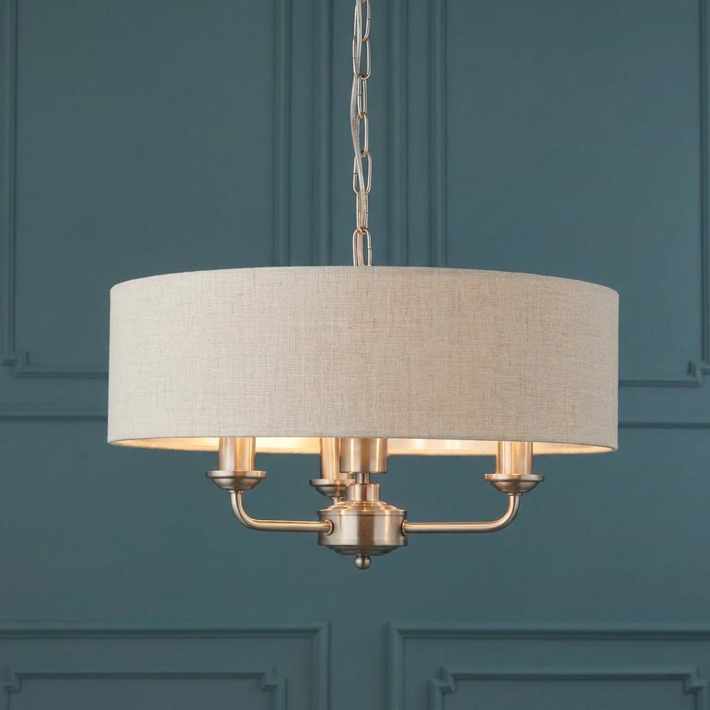 Three-light pendant with a natural linen drum shade and brushed chrome frame against a blue paneled wall.