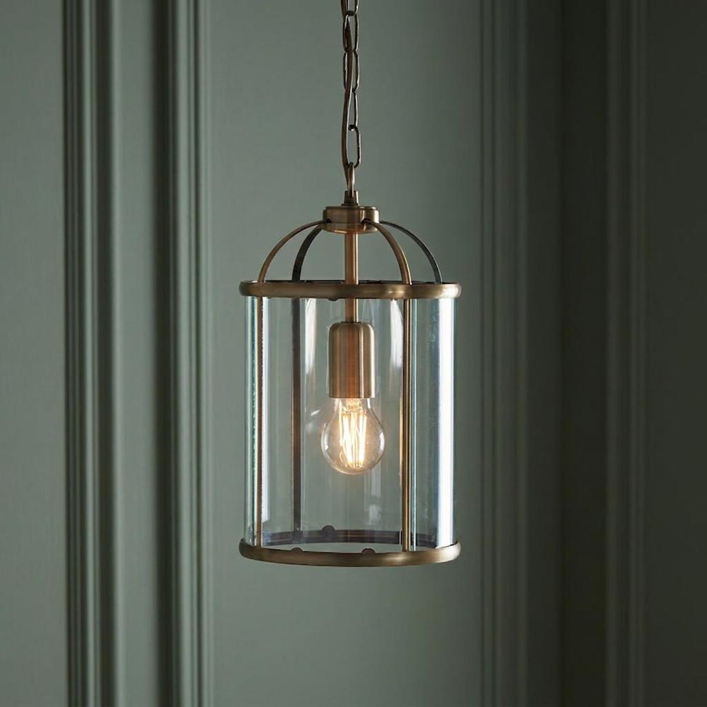 Antique brass lantern pendant light with clear glass and a glowing bulb, hanging against a paneled wall.