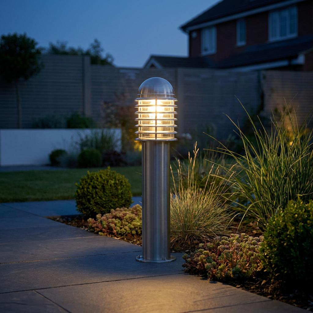 Illuminated stainless steel louvred bollard light standing next to a stone garden path at dusk.