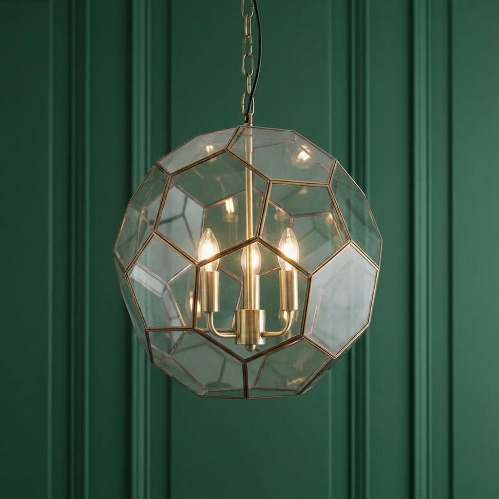 Geometric brass and glass globe pendant light with three glowing bulbs against a dark green paneled wall.