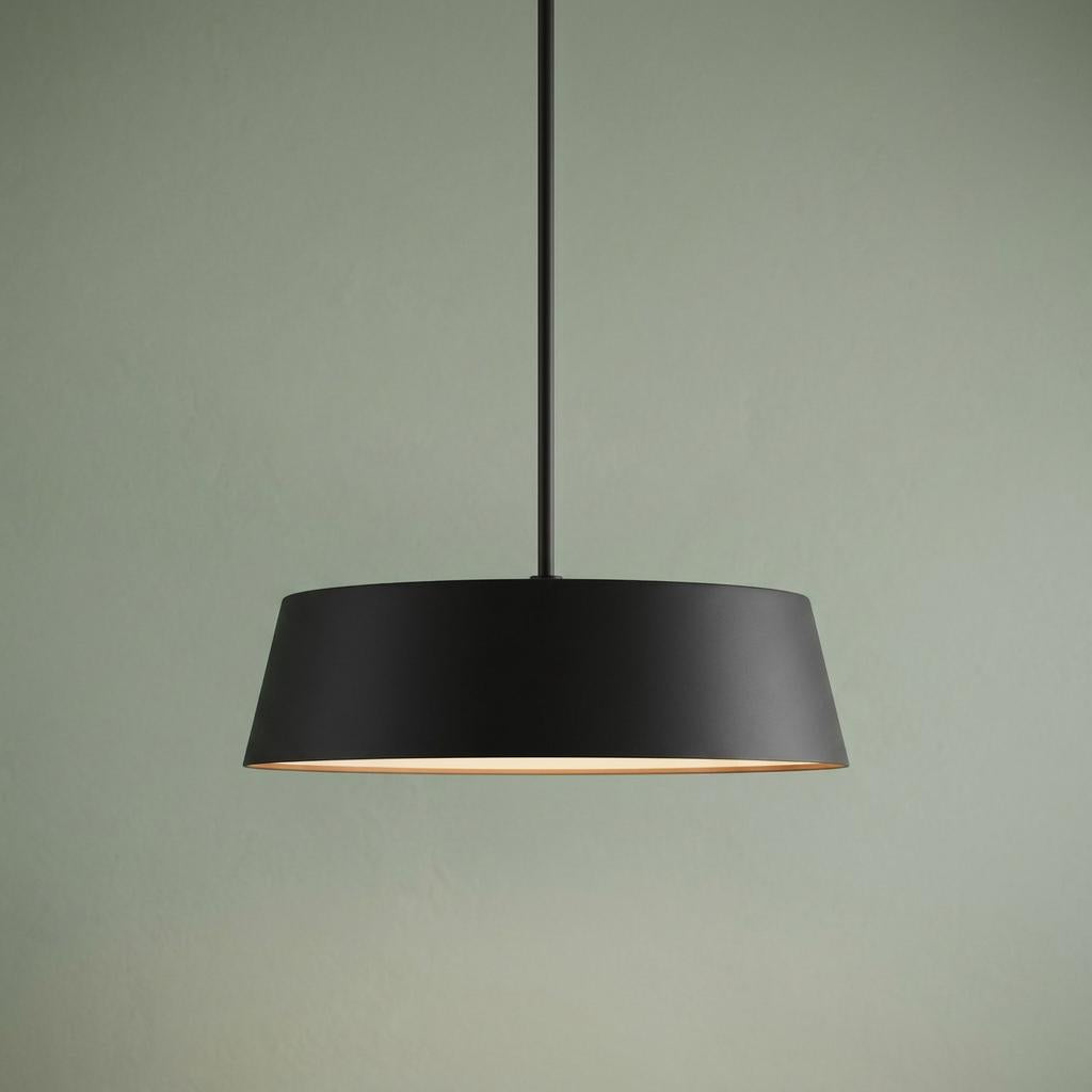 Modern black drum pendant light with a tapered shade hanging against a pale green wall, glowing with warm light.