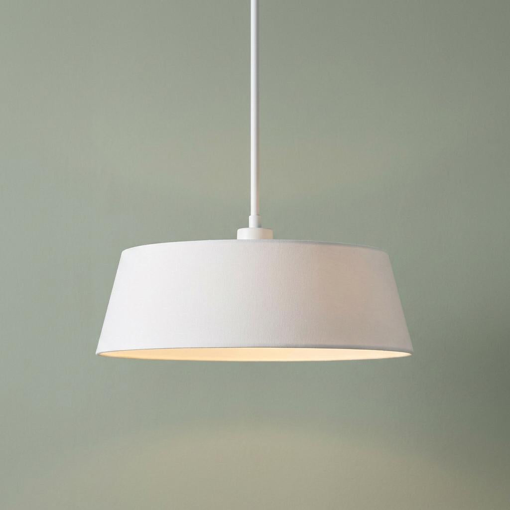 White tapered drum pendant light with a warm glow hanging against a sage green wall.
