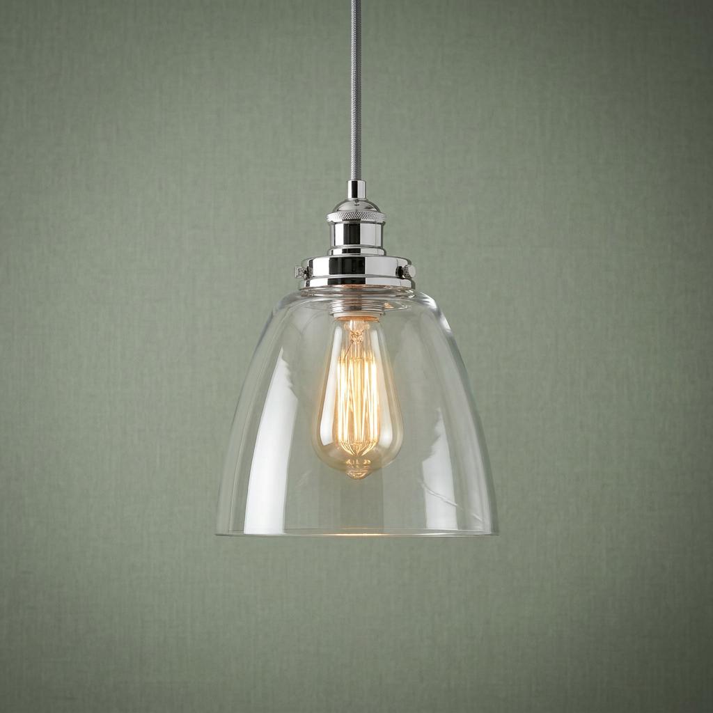 Polished nickel pendant light with a clear bell-shaped glass shade and warm Edison bulb against a green wall.