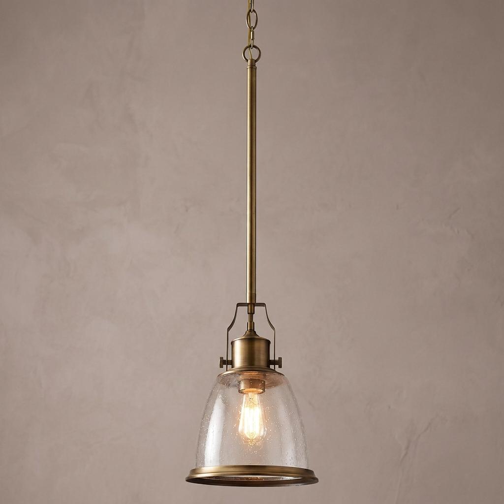 Aged brass ceiling pendant with clear seeded glass bell shade and exposed bulb against a neutral textured wall.