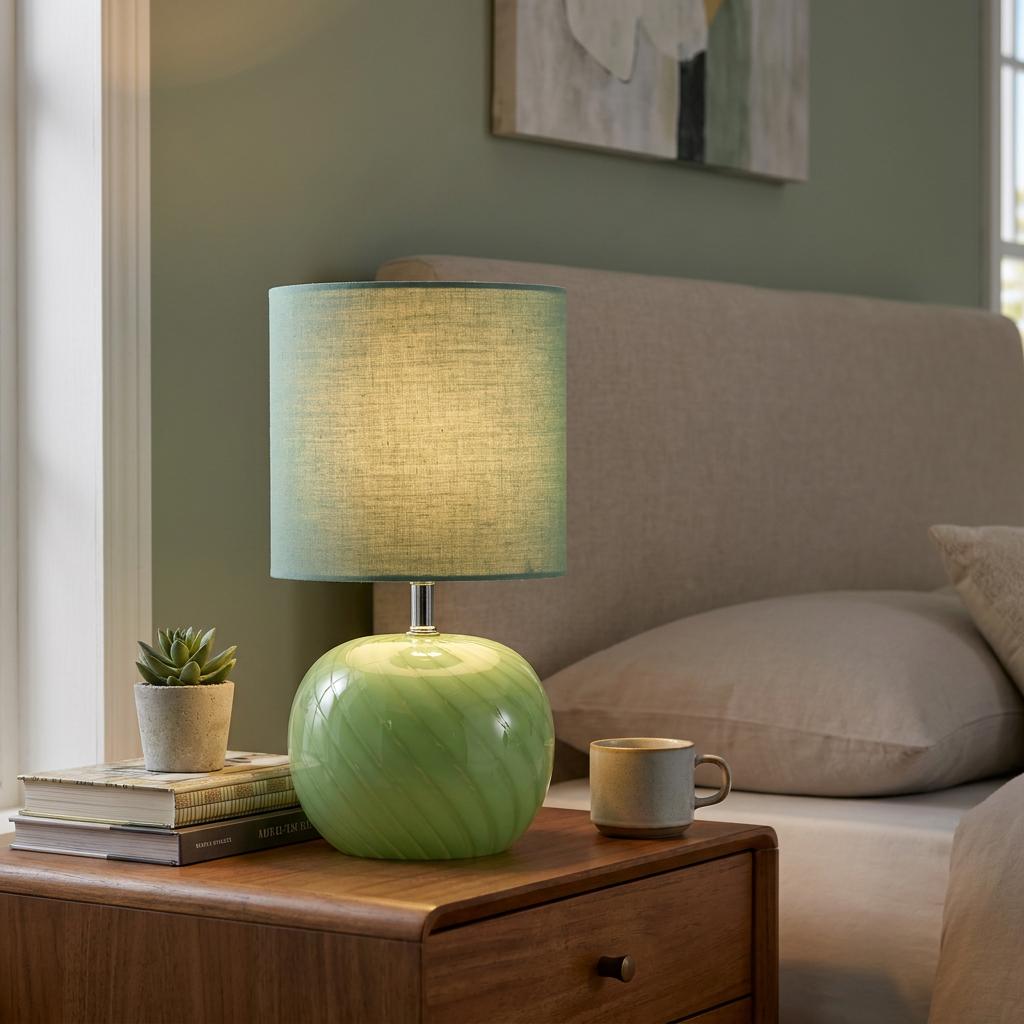 A green glass table lamp with a matching linen shade sits illuminated on a wooden bedside table.