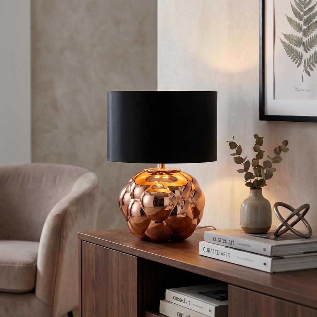 Fizz copper glass table lamp with a black faux silk shade sits on a wooden sideboard in a stylish living room.