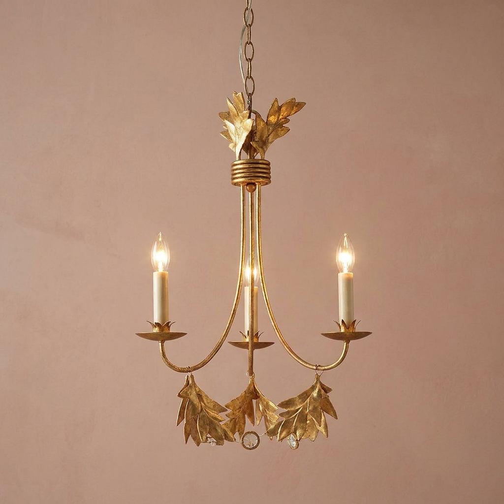 Three-light gilded gold chandelier with olive leaf motifs and lit candle bulbs against a plain pink wall.