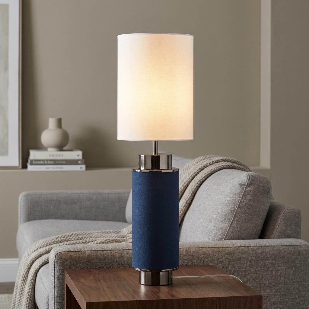 A blue linen and black nickel table lamp with a white shade on a wooden side table next to a grey sofa.